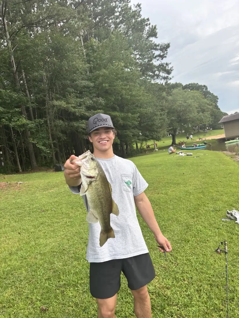 AJ Phillips standing on a grass bank by a lake holding up a largemouth bass he has just caught