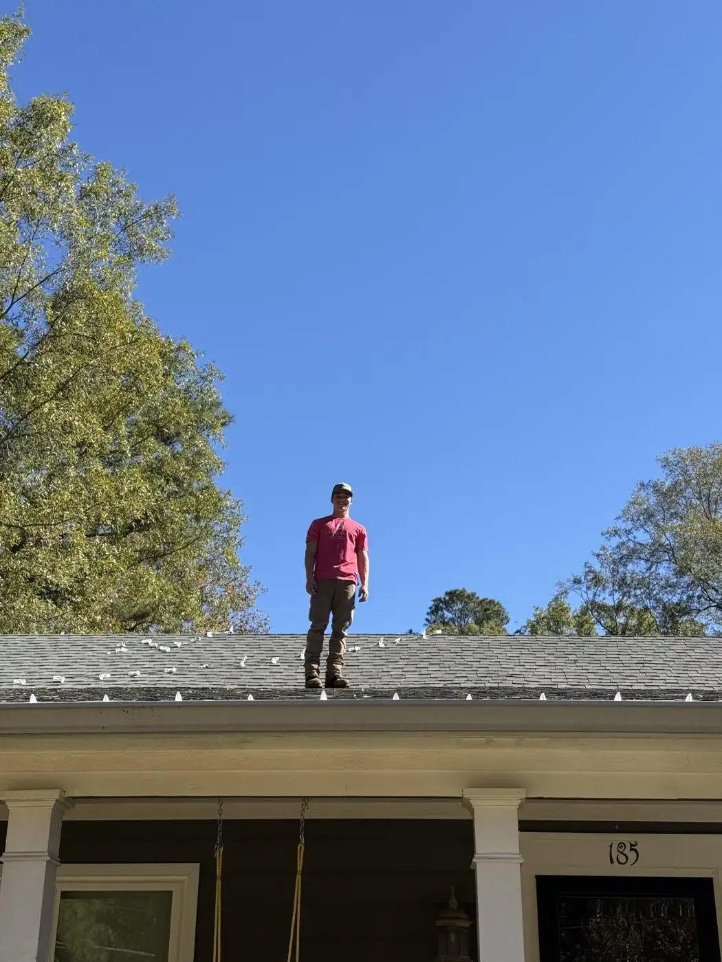 AJ Phillips standing on the ridge of a shingle roof against a clear blue sky, with a finished run of light clips already spaced along the edge below him