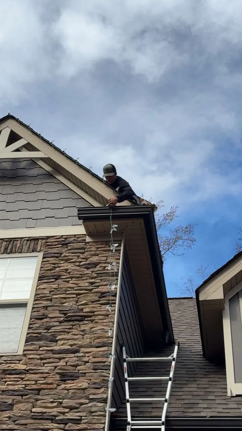 View up a ladder to a stone-and-siding gable where AJ Phillips is feeding a strand of lights down the corner of the gutter line