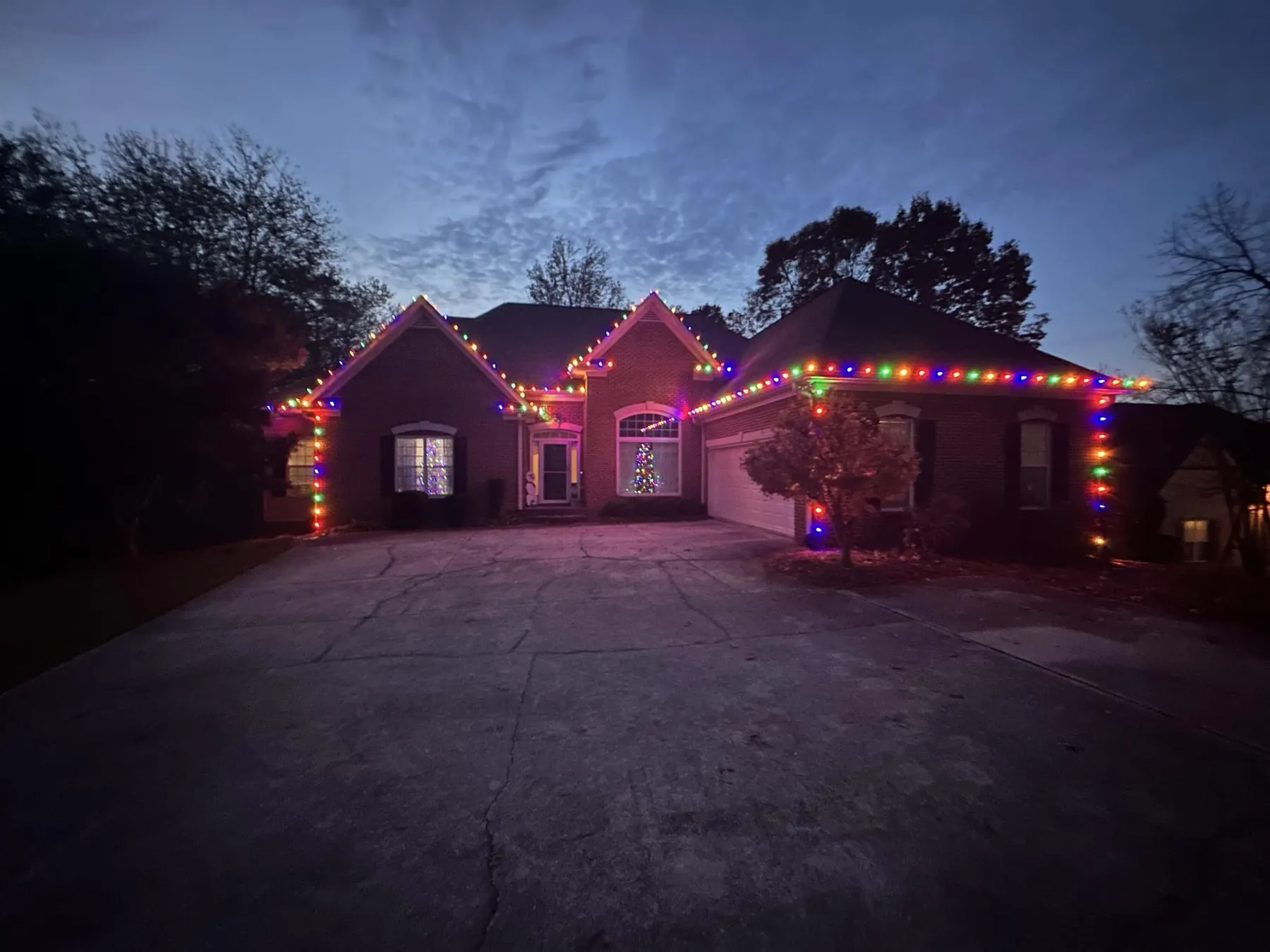 Brick home at dusk with multicoloured C9 bulbs along every roofline and gable, a lit tree visible in the front window and the driveway in the foreground