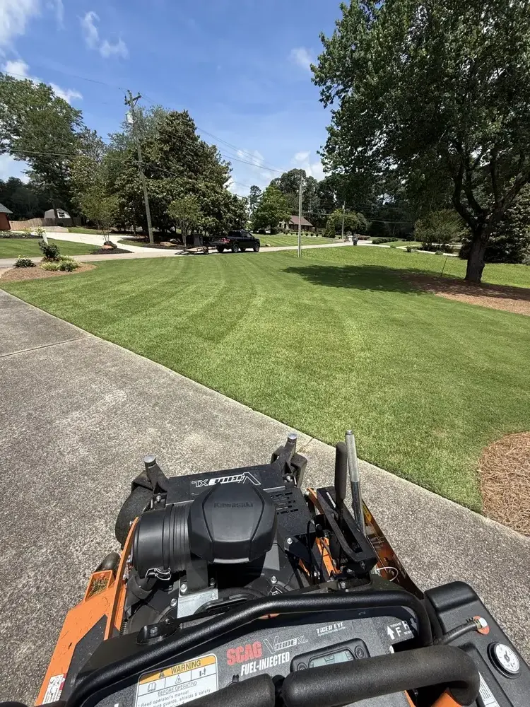 Wide front lawn cut into clean light and dark stripes, viewed over the handlebars of an orange Scag V-Ride XL stand-on mower parked on the driveway